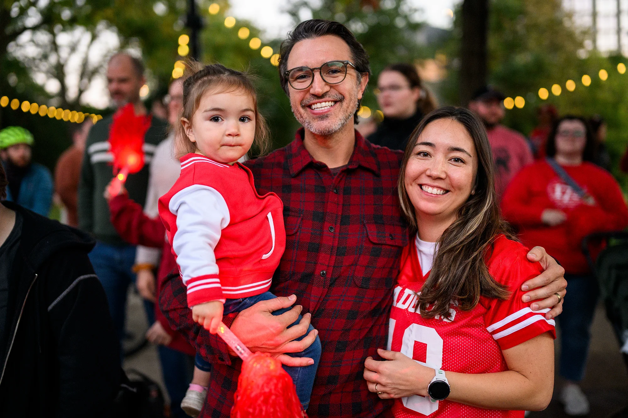 A smiling man holding a child in a mini red letter jacket stands with a woman in a red 'Badgers' jersey surrounded by a festive crowd.