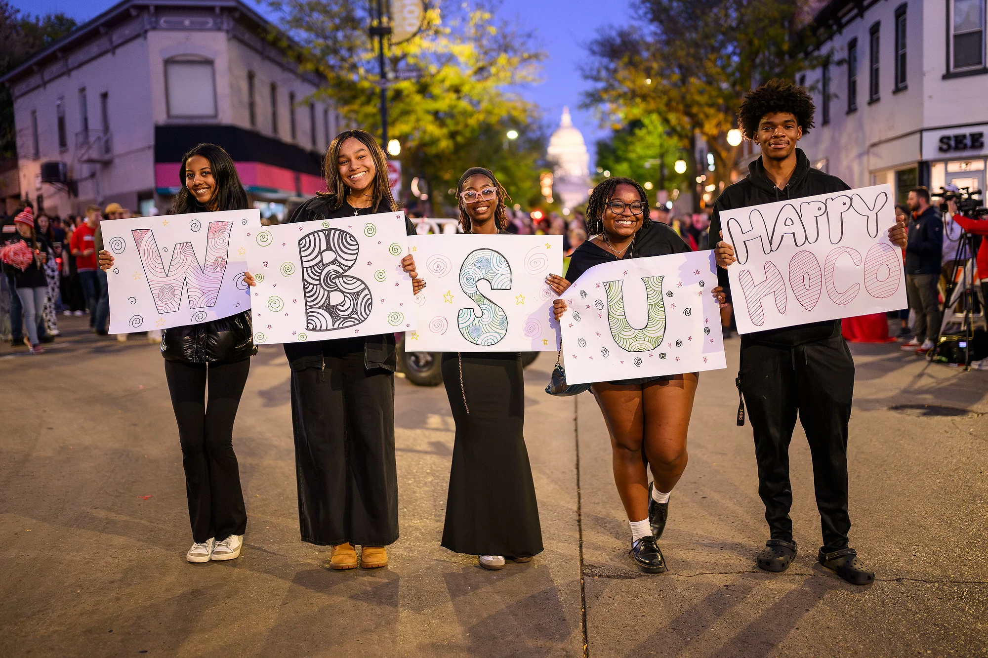 Five individuals hold colorful signs spelling 'WBSU HAPPY HOCO' while walking down State street in the Homecoming Parade. The capitol building glows in the background.