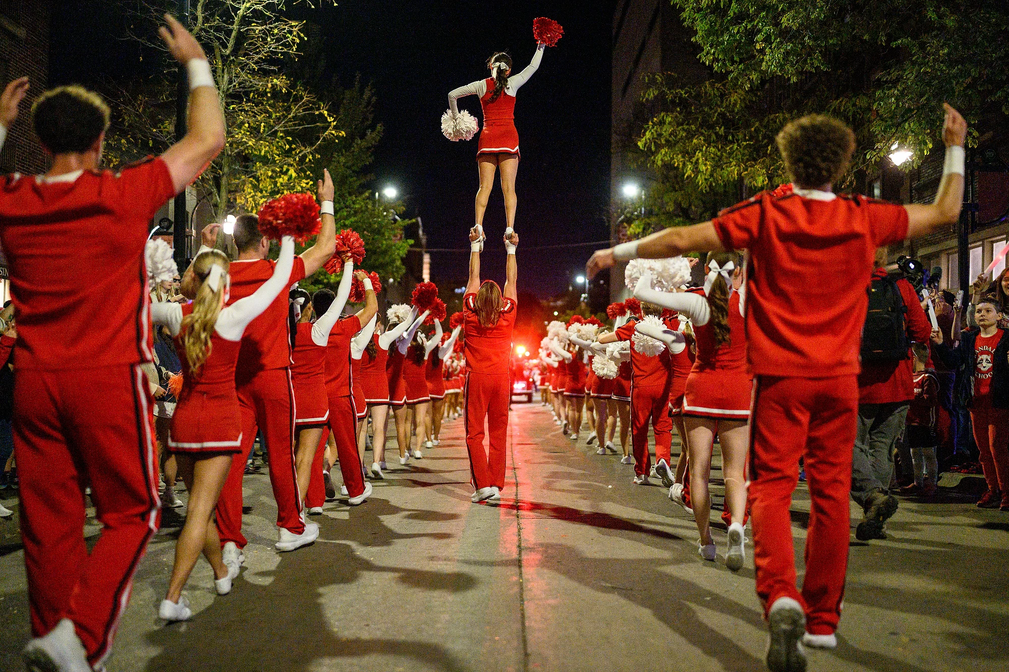 A cheerleader is lifted high into the air by her teammate during a choreographed nighttime performance on State Street, surrounded by spectators.