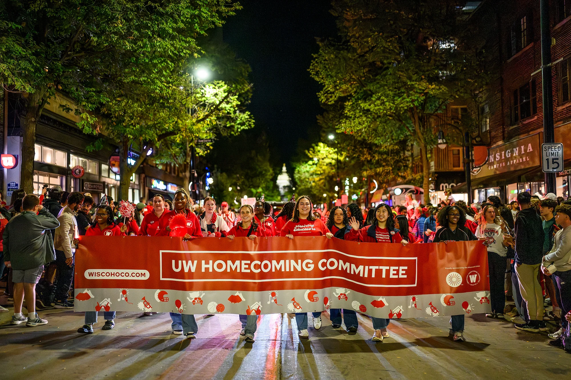 Marchers in red shirts carry a large 'UW Homecoming Committee' banner down State street, with spectators lining the route.