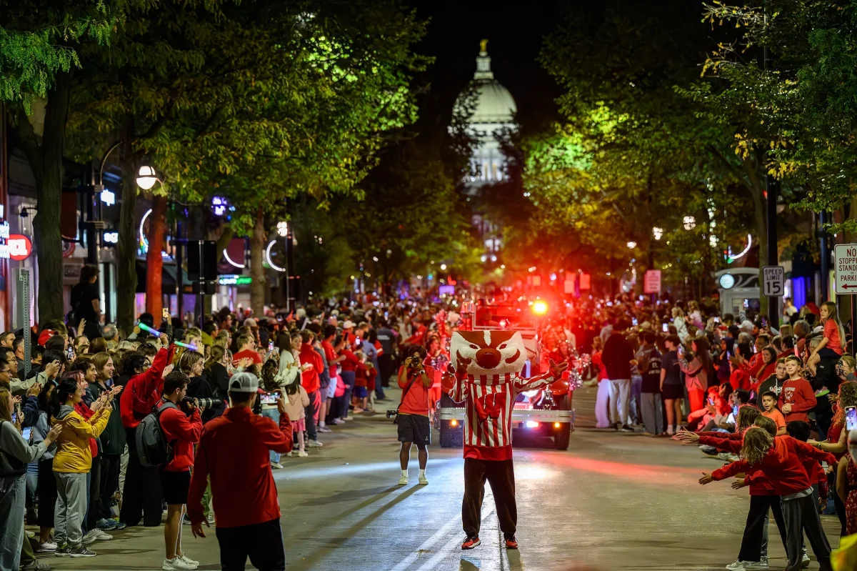 Bucky Badger leads the nighttime homecoming parade down State Street with the Wisconsin capitol building illuminated in the background as fans and alumni line the sidewalks and cheer