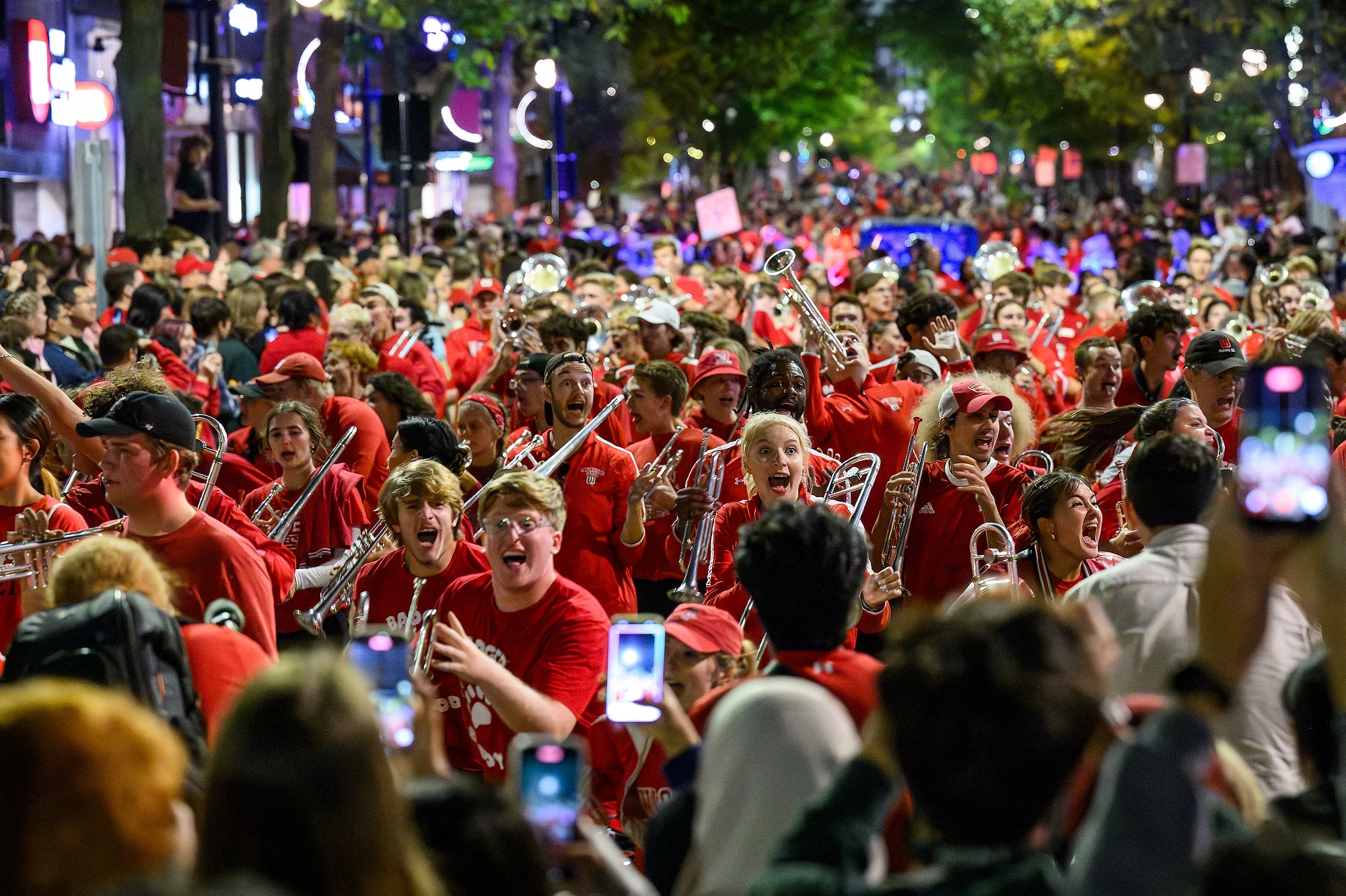 A spirited marching band dressed in red fills part of State street with energy during UW–Madison’s nighttime Homecoming parade, surrounded by cheering spectators.
