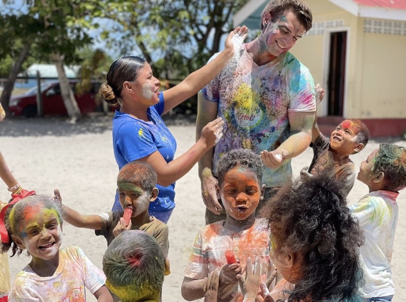 A joyful group of children and adults celebrate outdoors, playfully tossing vibrant colored powders in a festive, Holi-inspired gathering.