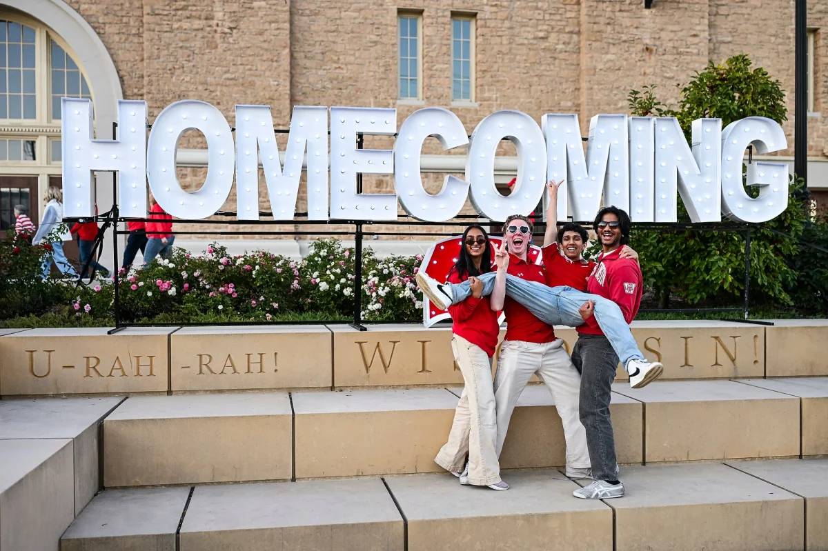 Four friends all wearing red pose in front of a large "Homecoming" sign in Alumni Park