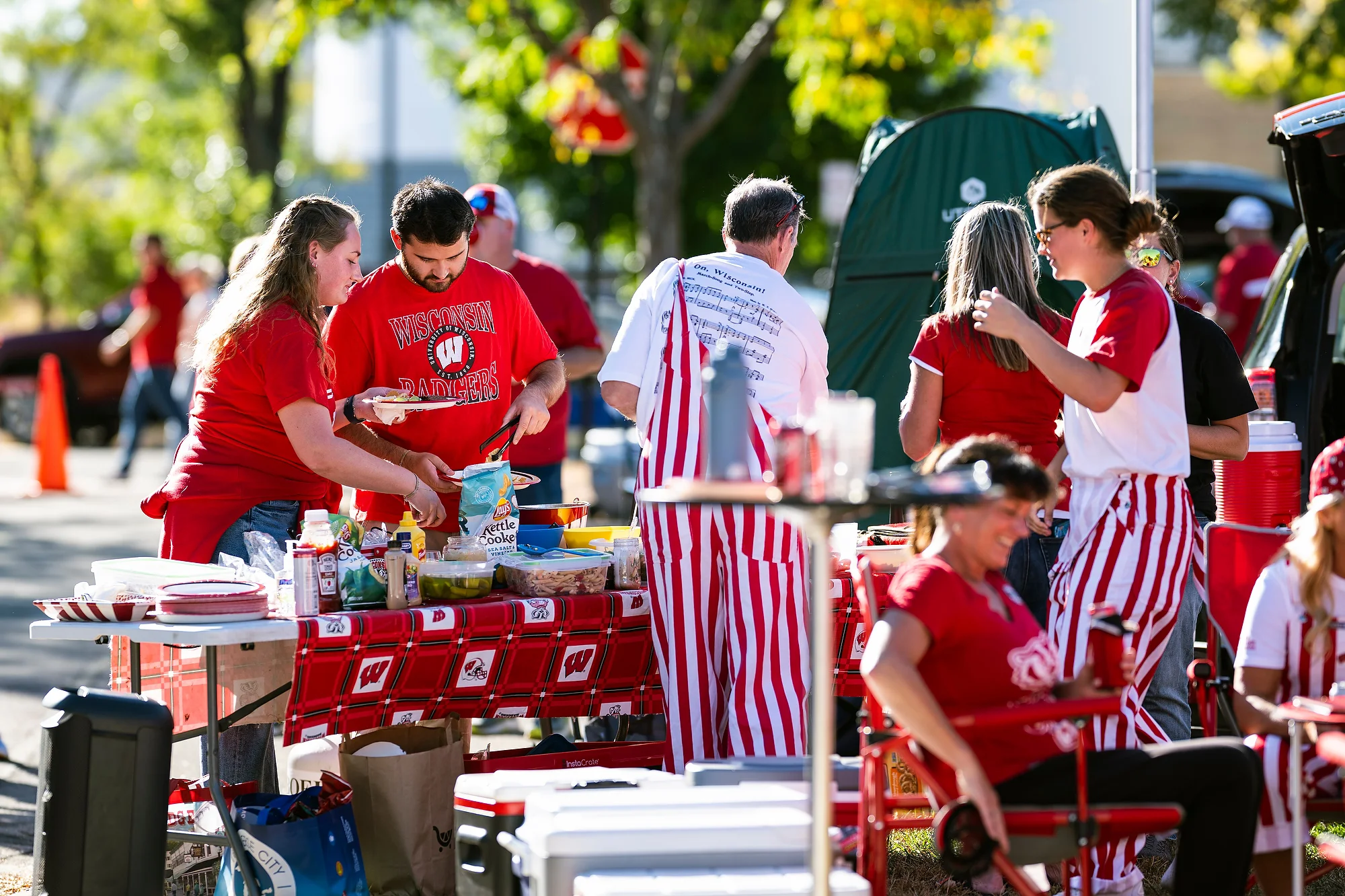 Badger fans wearing red and white striped overalls enjoy food and beverages outdoors.