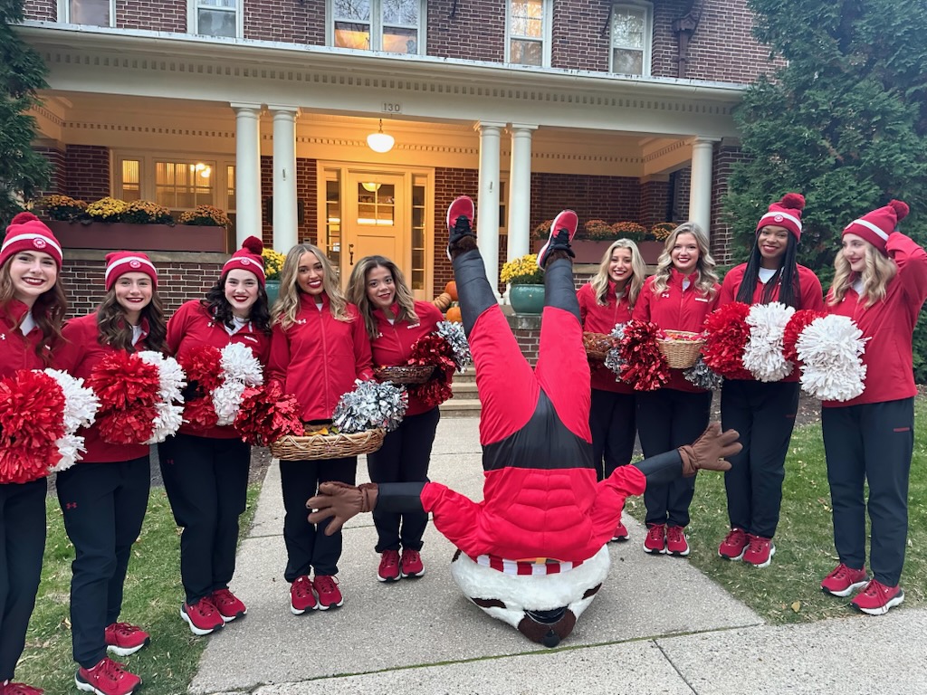 A group of students with their spirit pom poms stand in a line while Bucky Badger does a handstand.