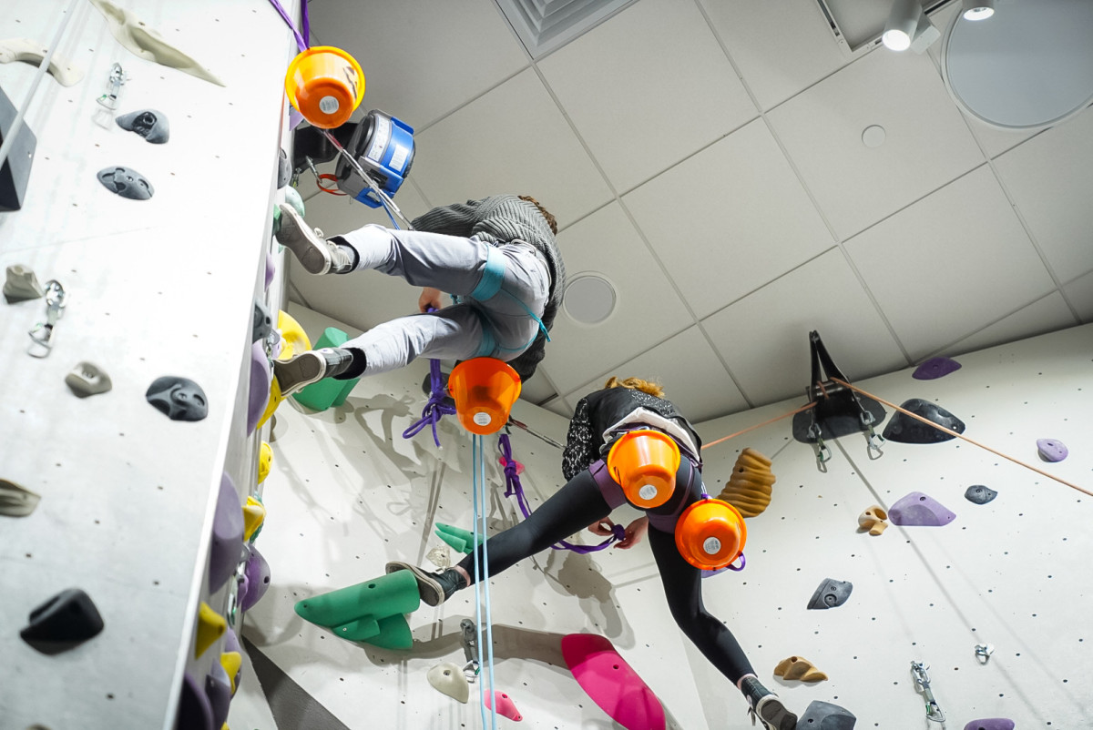 Two students scale a climbing wall with Halloween candy buckets strapped to their harnesses.