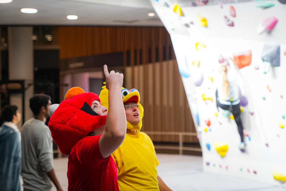 Two students dressed in an Elmo and Big Bird costume stand at a climbing wall and discuss their climbing approach.