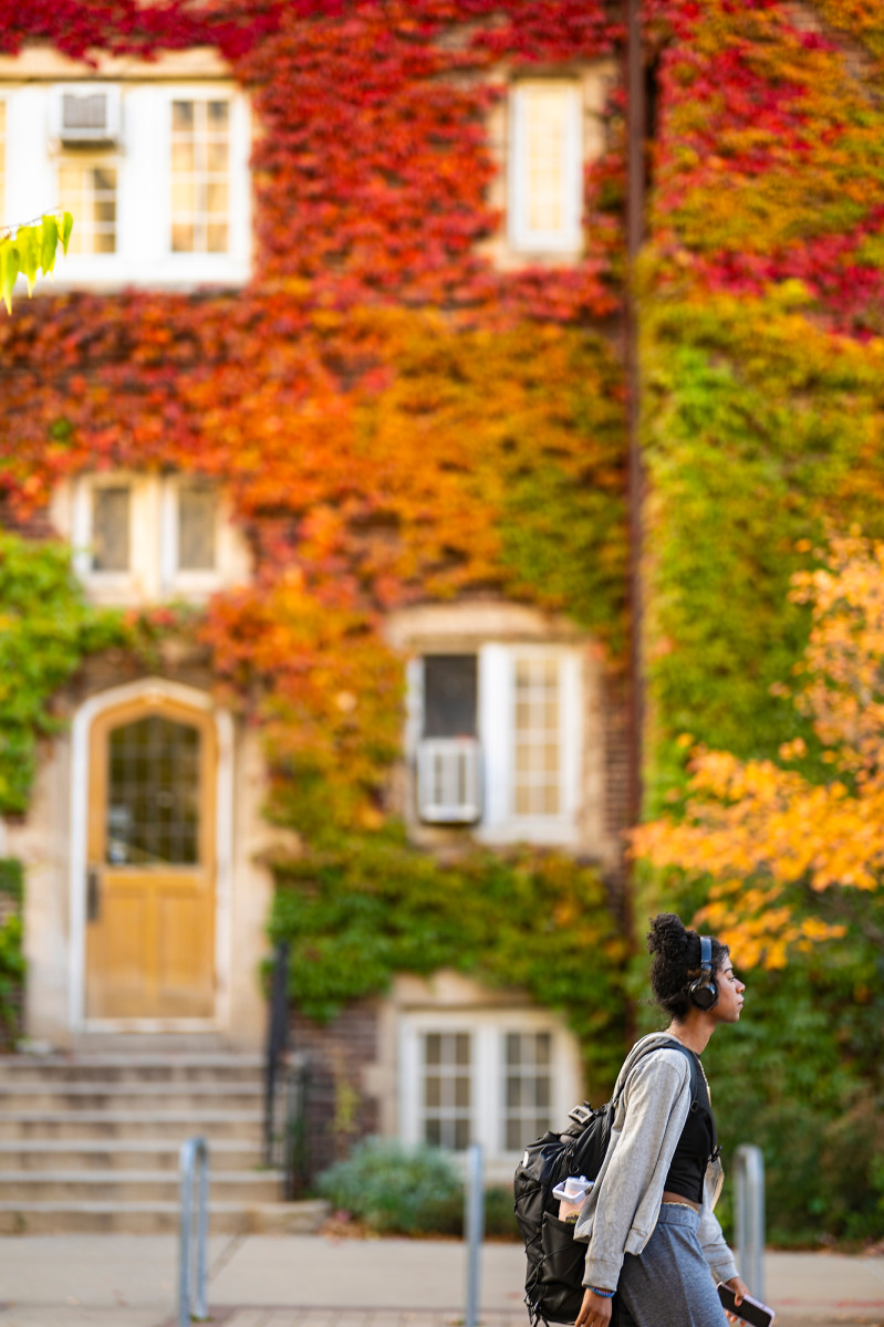 A person with headphones walks past a building covered in colorful fall leaves