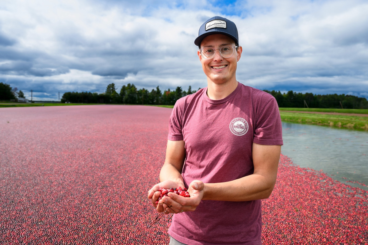 A man stands in a cranberry bog at harvest time. He smiles while holding up a handful of cranberries.