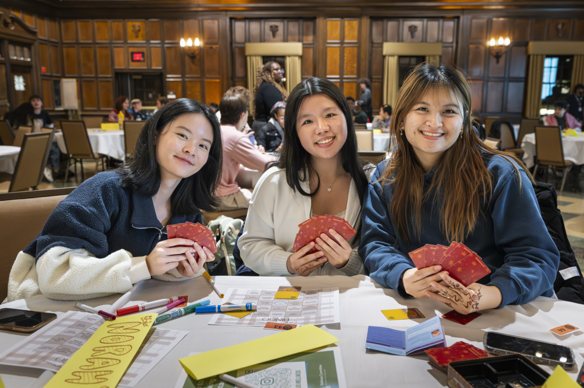 Three students, each holding playing cards in their hands, sit at a table and smile for the camera.