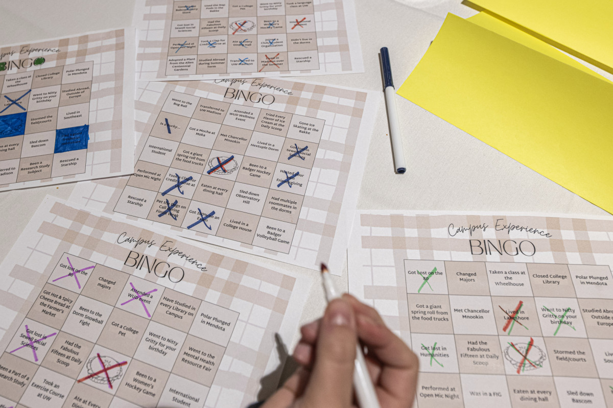 A close-up photo of several marked BINGO sheets on a table. A hand holding a marker appears, ready to mark off another space on one of the cards.
