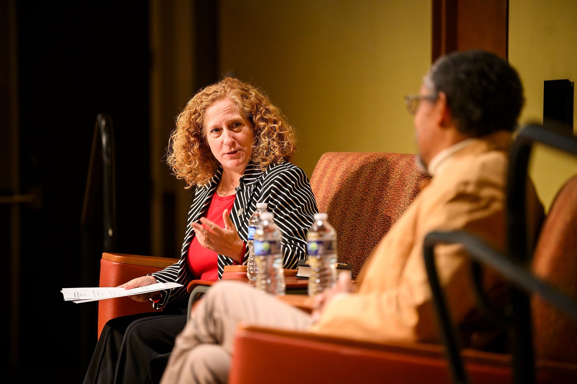 A woman and a man sit in chairs on a stage talking, the woman leaning forward to ask a question.