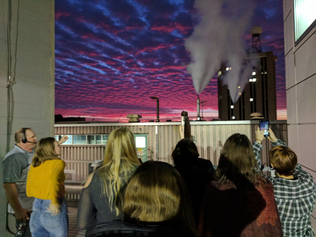 A group of college students are in a building looking out a huge window that shows a sunset.