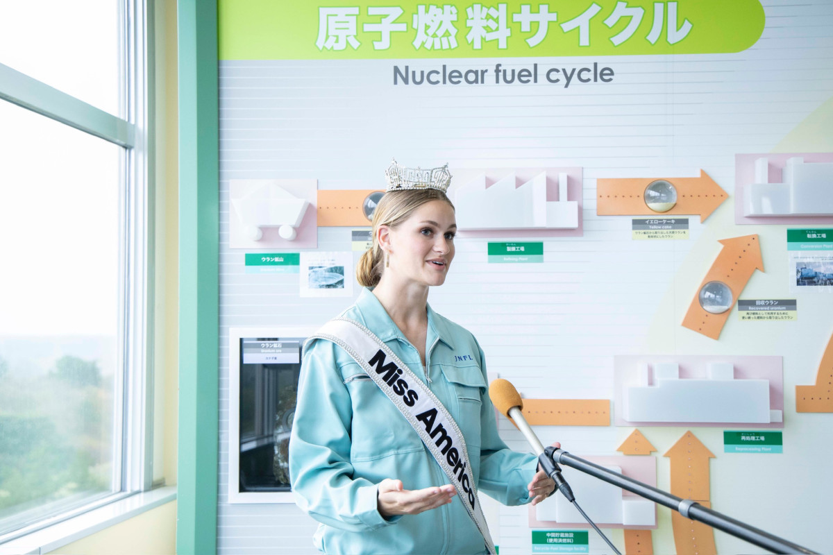 A woman in a jumpsuit wearing a tiara and a sash that says Miss America stands and speaks at a microphone.