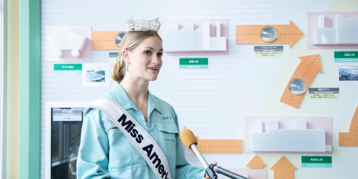 A woman in a jumpsuit wearing a tiara and a sash that says Miss America stands and speaks at a microphone.