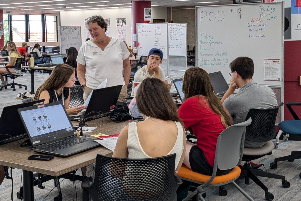 A woman stands and addresses several students seated at a table, looking at laptops.