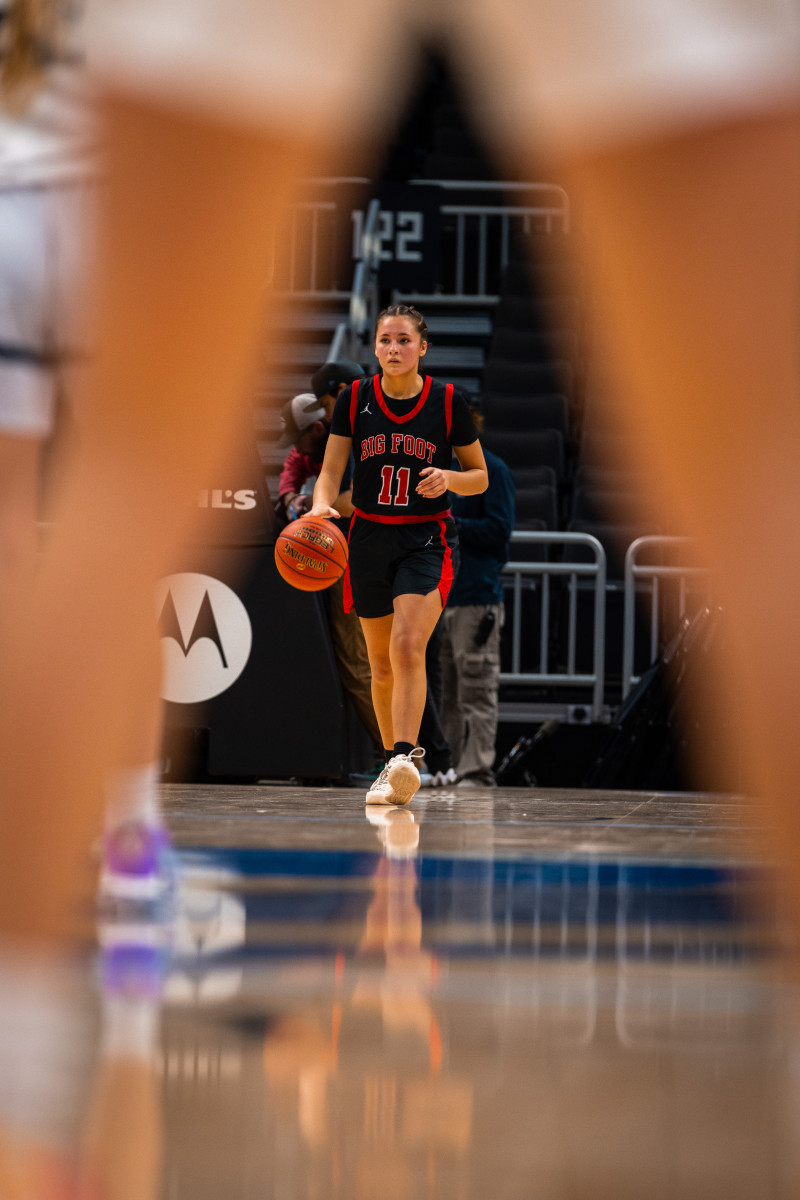 Through the legs of an opponent, a young woman is seen on the basketball court dribbling a basketball.