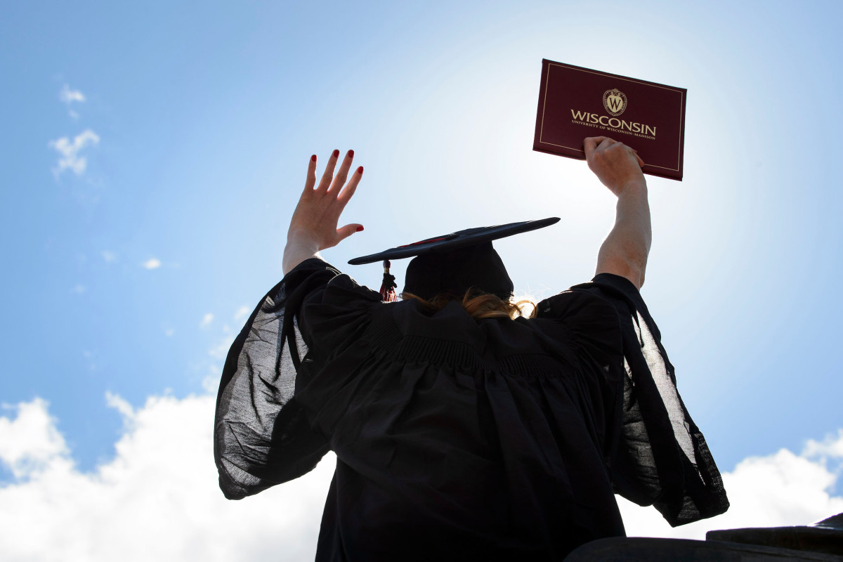 A student wears a graduation robe and cap and lifts her diploma up in the air.