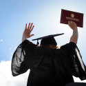 A student wears a graduation robe and cap and lifts her diploma up in the air.