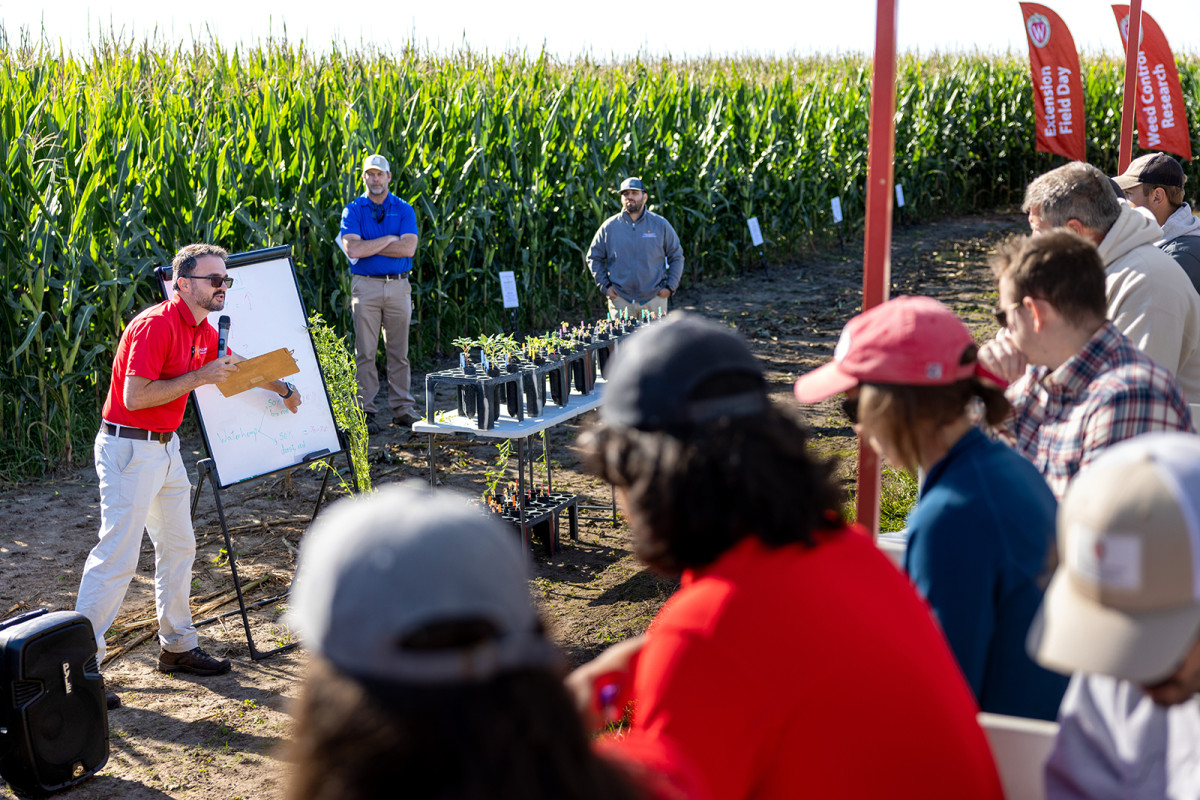 Rodrigo Werle stands in front of corn field and speaks into a microphone in front of a crowd of people