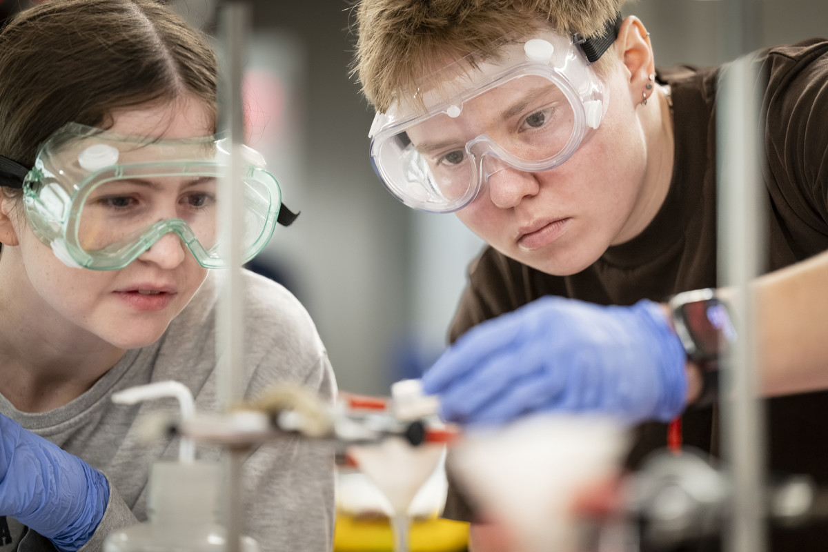 Two students with safety goggles pouring a substance into a funnel