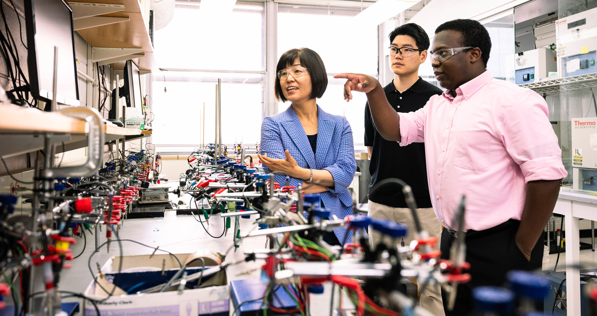 Three people looking at a monitor next to a bench of scientific equipment