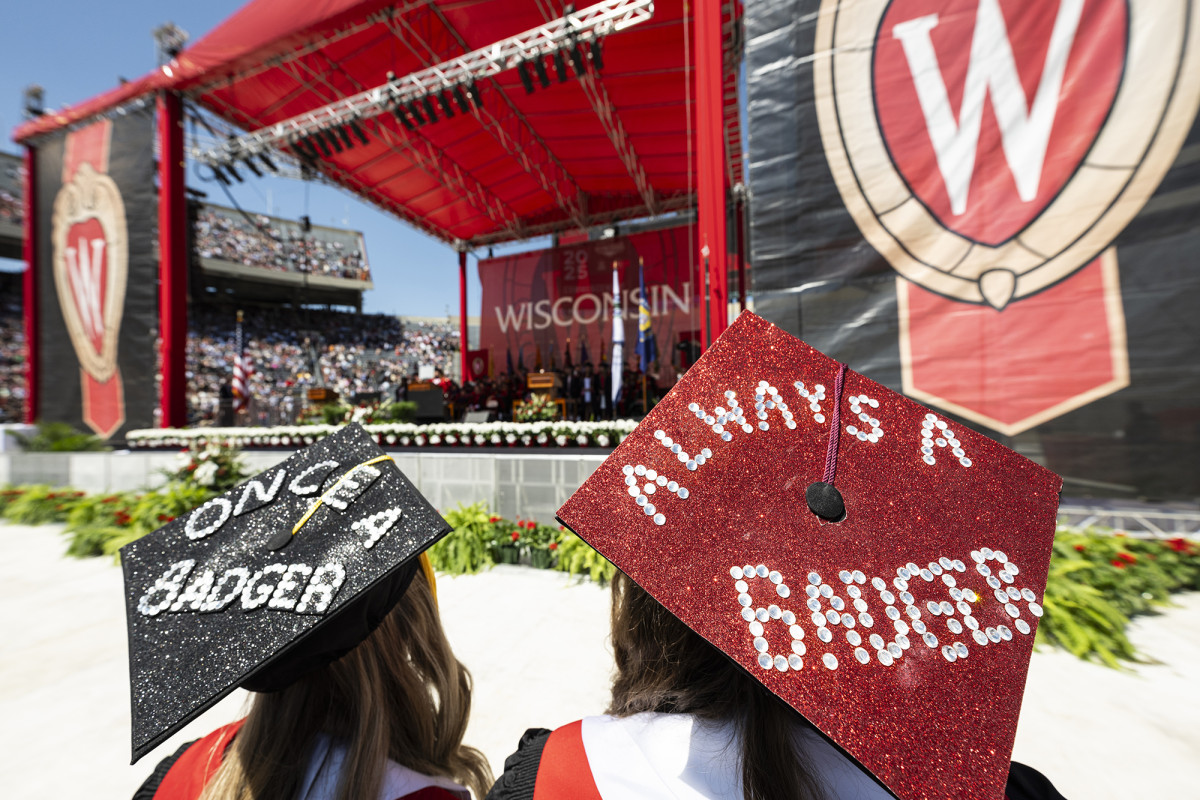 A view of the back of two graduation caps being worn by two people with a stage in the background