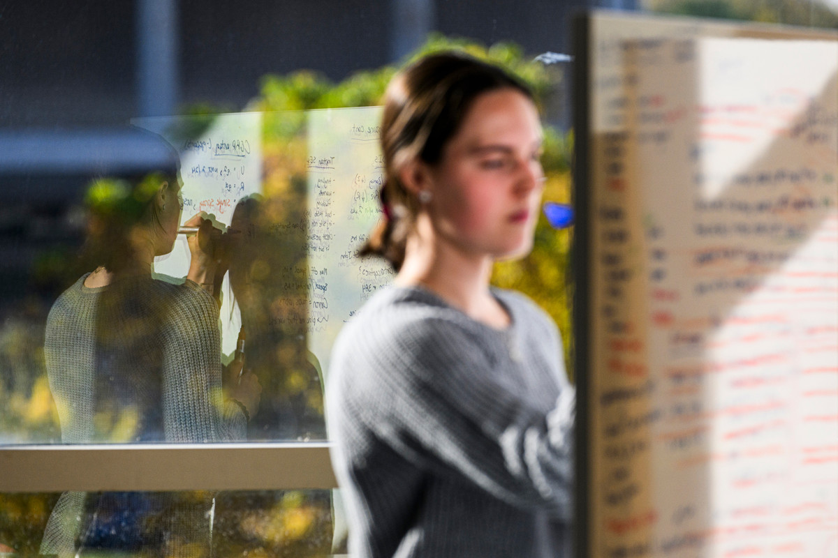 A person writing on a white board with their reflection on the window behind them