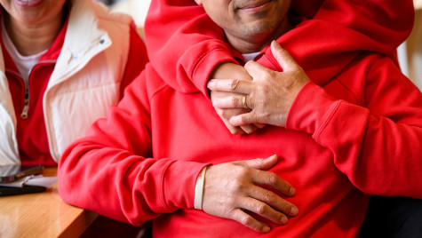 A person stands behind a seated man and wraps their arms around him.