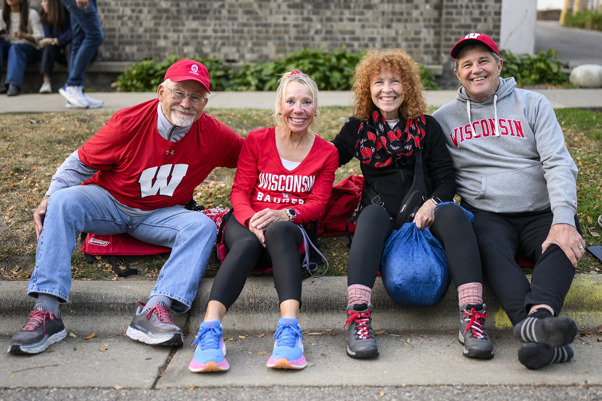 Four people sitting on a curb wearing UW–Madison attire smiling
