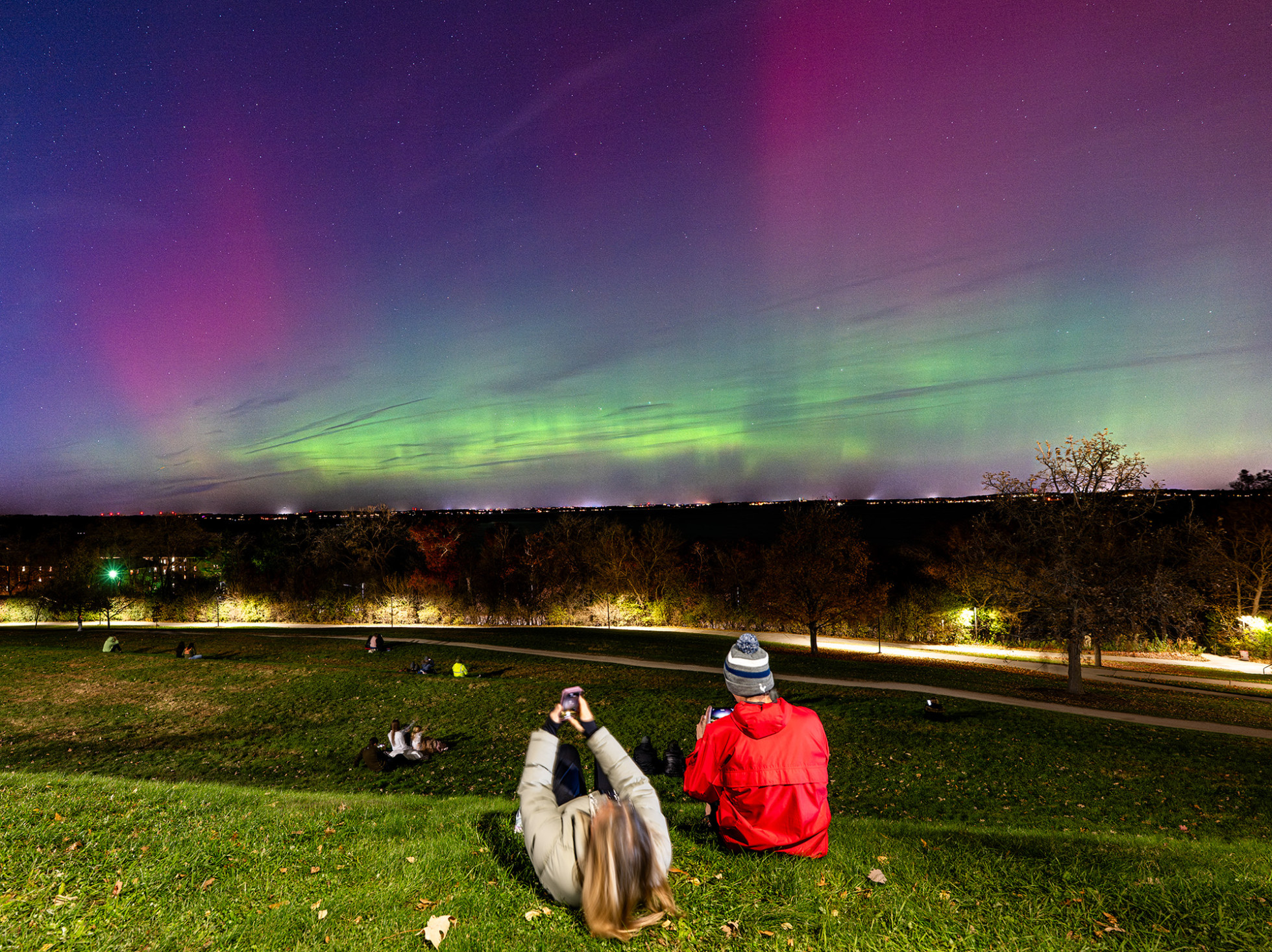 Several groups of people sitting on hillside while observing the Northern Lights in the night sky