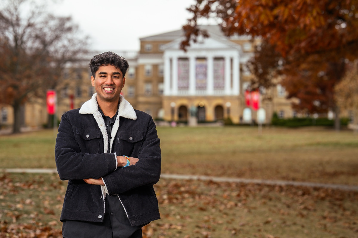 A student wearing a fall coat smiles and stands with his arms crossed. He's standing on Bascom Hill on an autumn day.