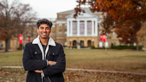 A student wearing a fall coat smiles and stands with his arms crossed. He's standing on Bascom Hill on an autumn day.