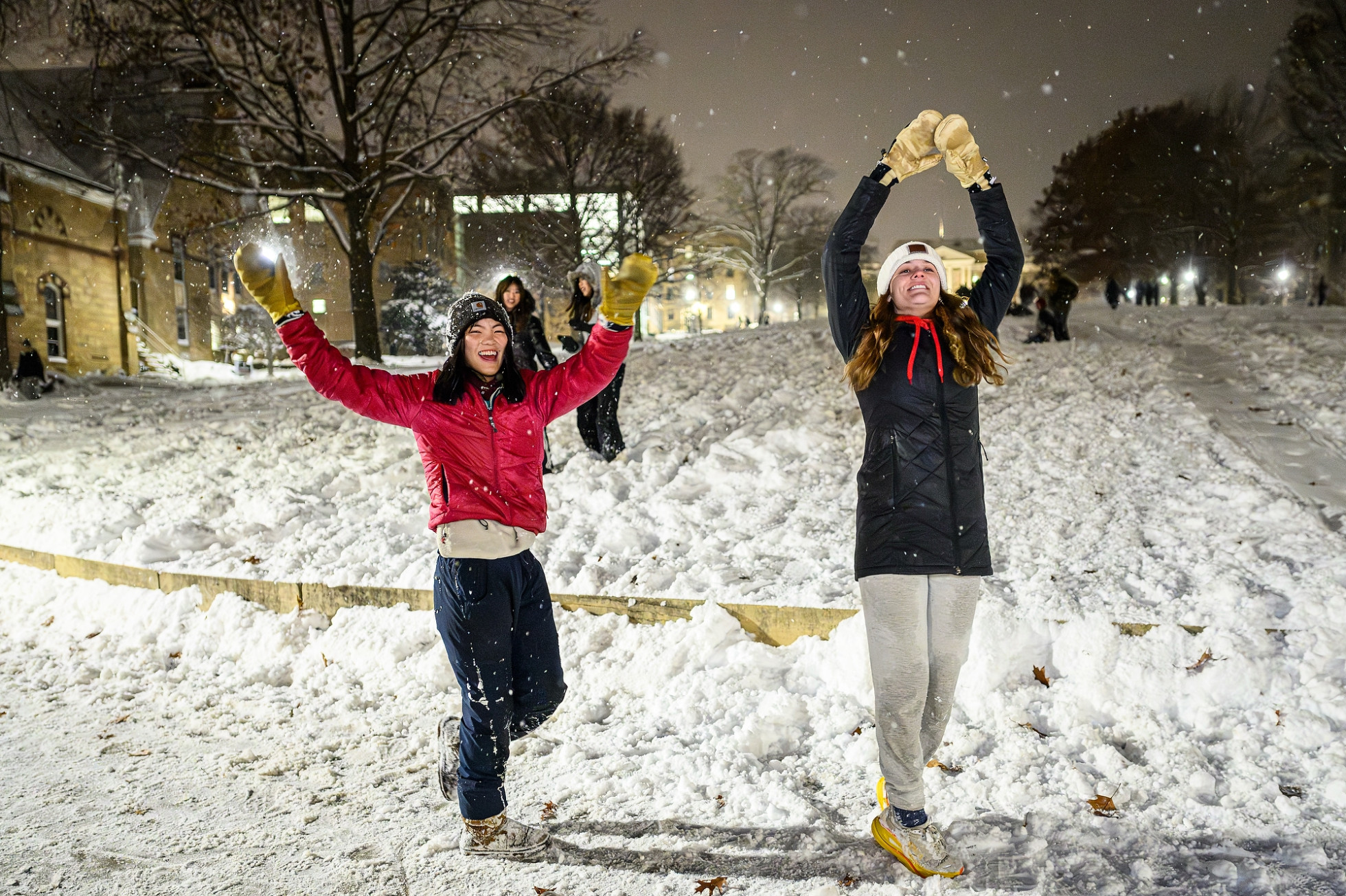 Two students wearing snow gloves and colorful jackets reaching up into the sky while smiling in the falling snow