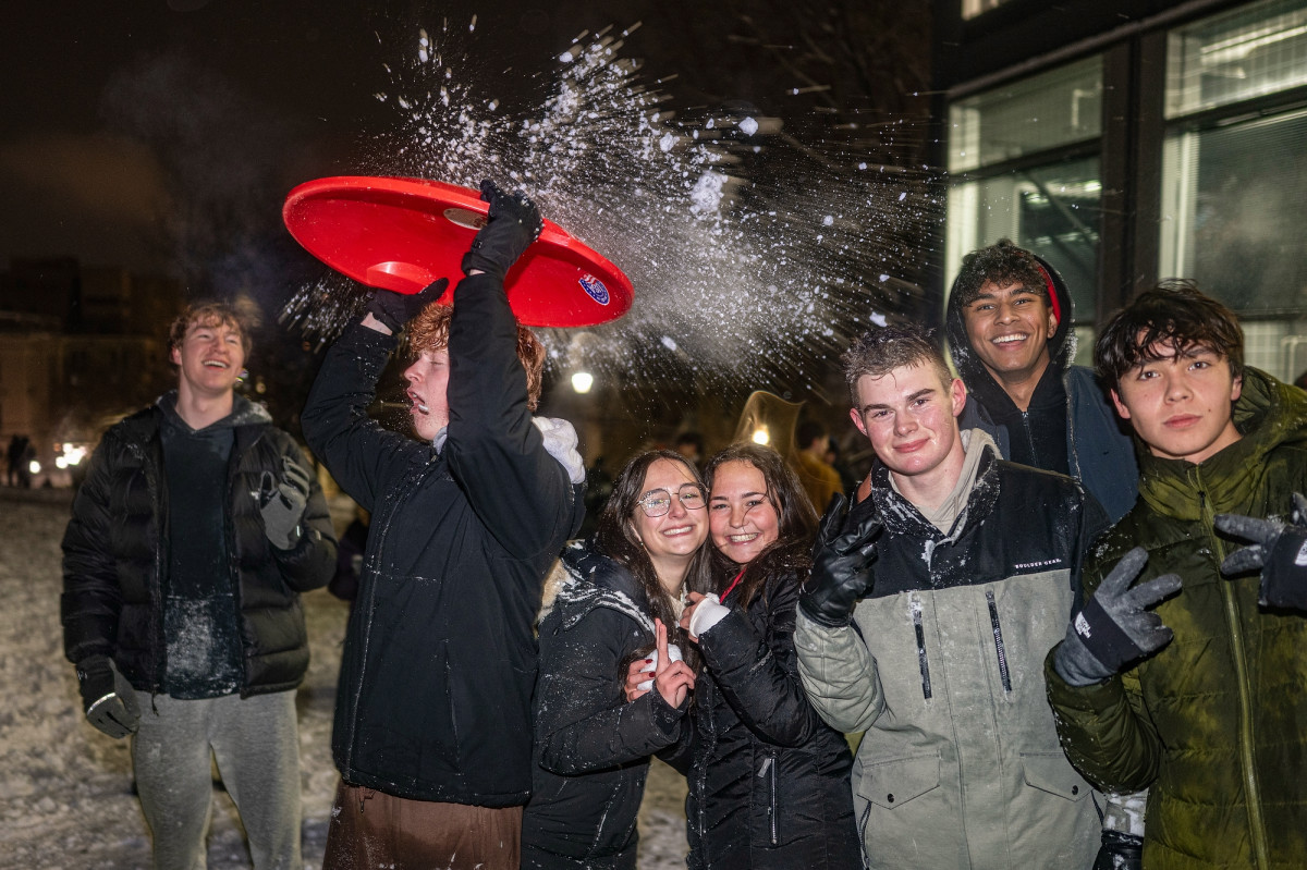 A student blocking snow with a shield sending it flying over students smiling for a group photo