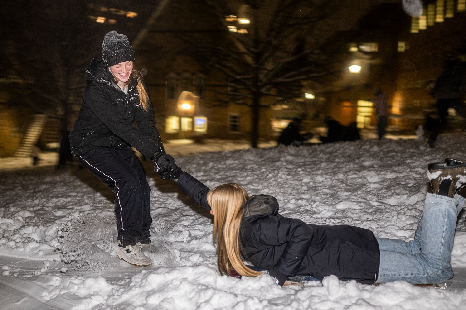 A students pulling another by the hand to try to get her sled moving on snowy Bascom Hill