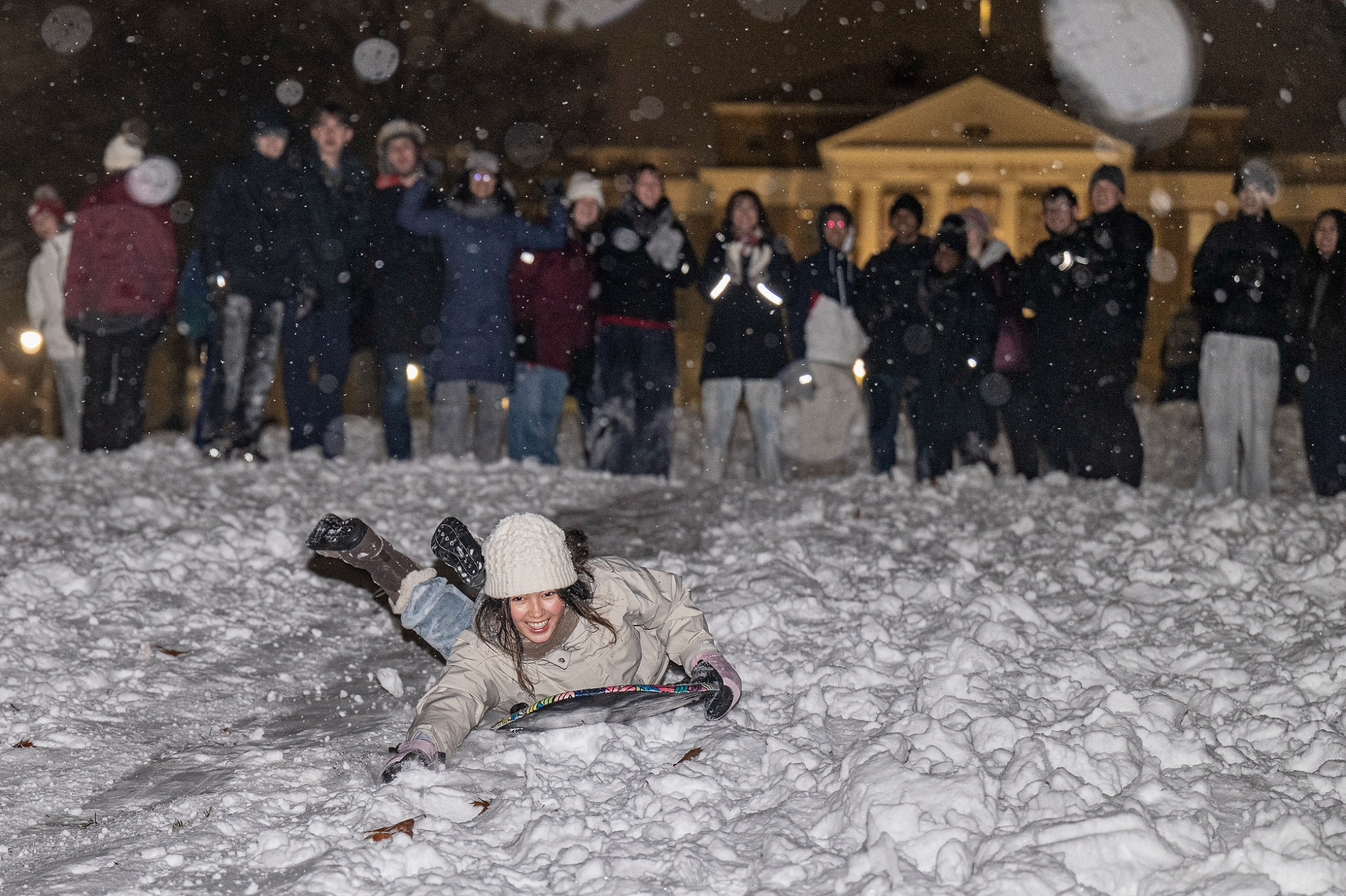 A student sledding down snowy Bascom Hill on her stomach while a group of students cheer her on