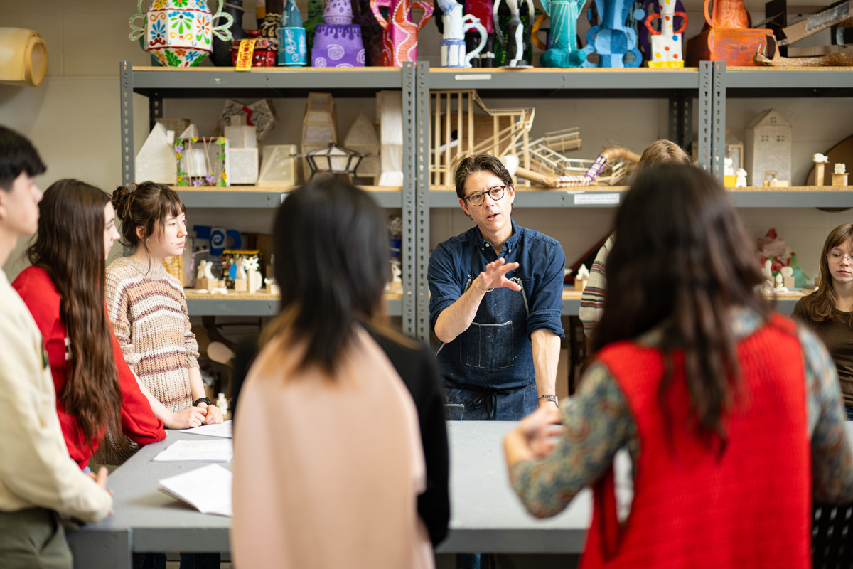 A professor talking to a group of students with artwork on shelves in the background