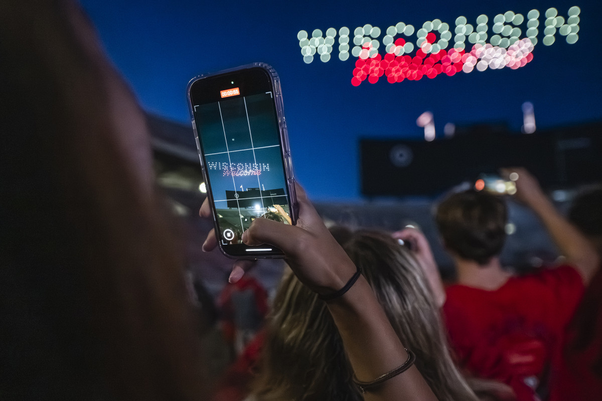 A person taking a photo with their phone of a drone light display reading "Wisconsin Welcome"