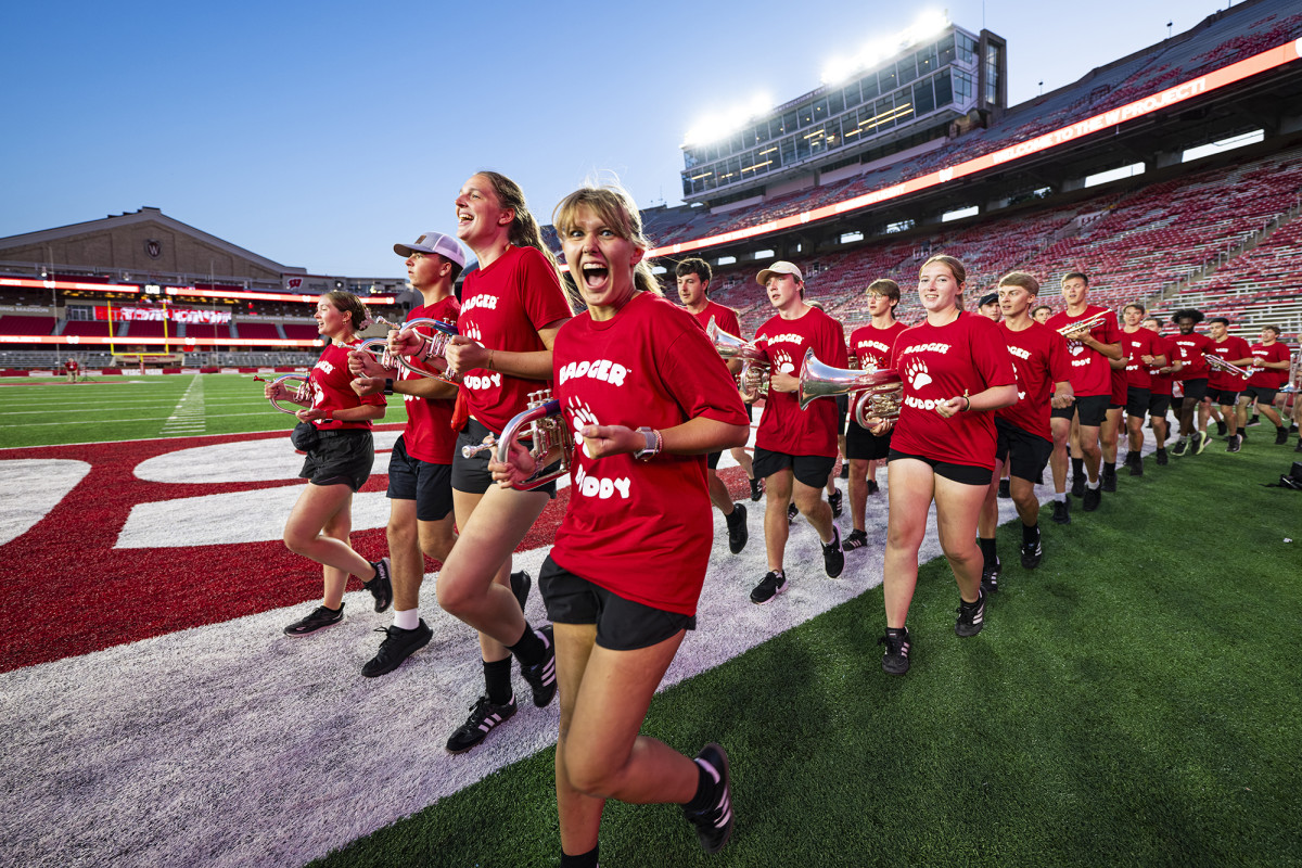 Large group of band members in red shirts jogging across a football field with their instruments