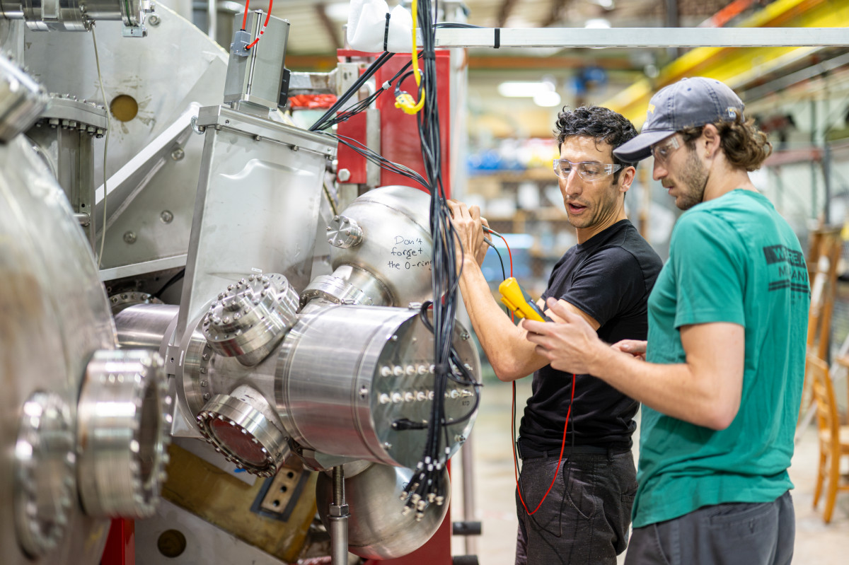 Two people working with large stainless steel equipment and wearing safety goggles