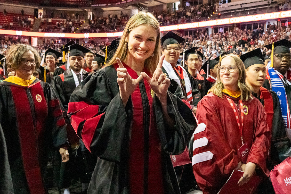 Keynote speaker Grace Vanderhei smiles and holds up her fingers in a W shape as she walks through the crowd of graduates.