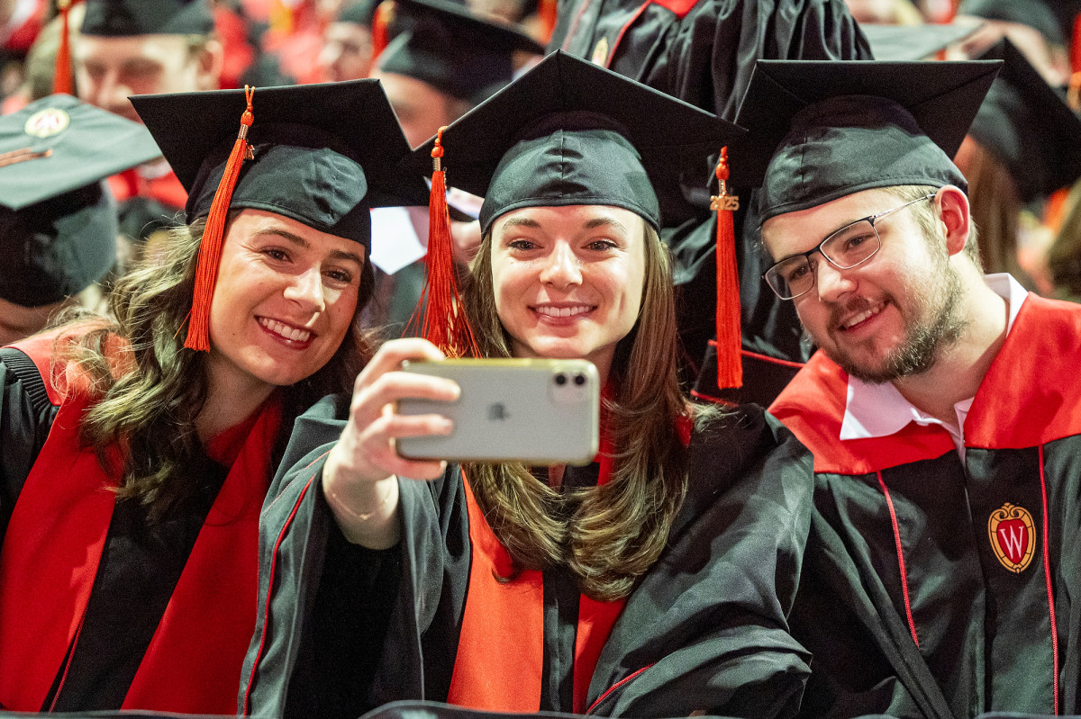 Three graduates in academic regalia lean together to take a selfie.