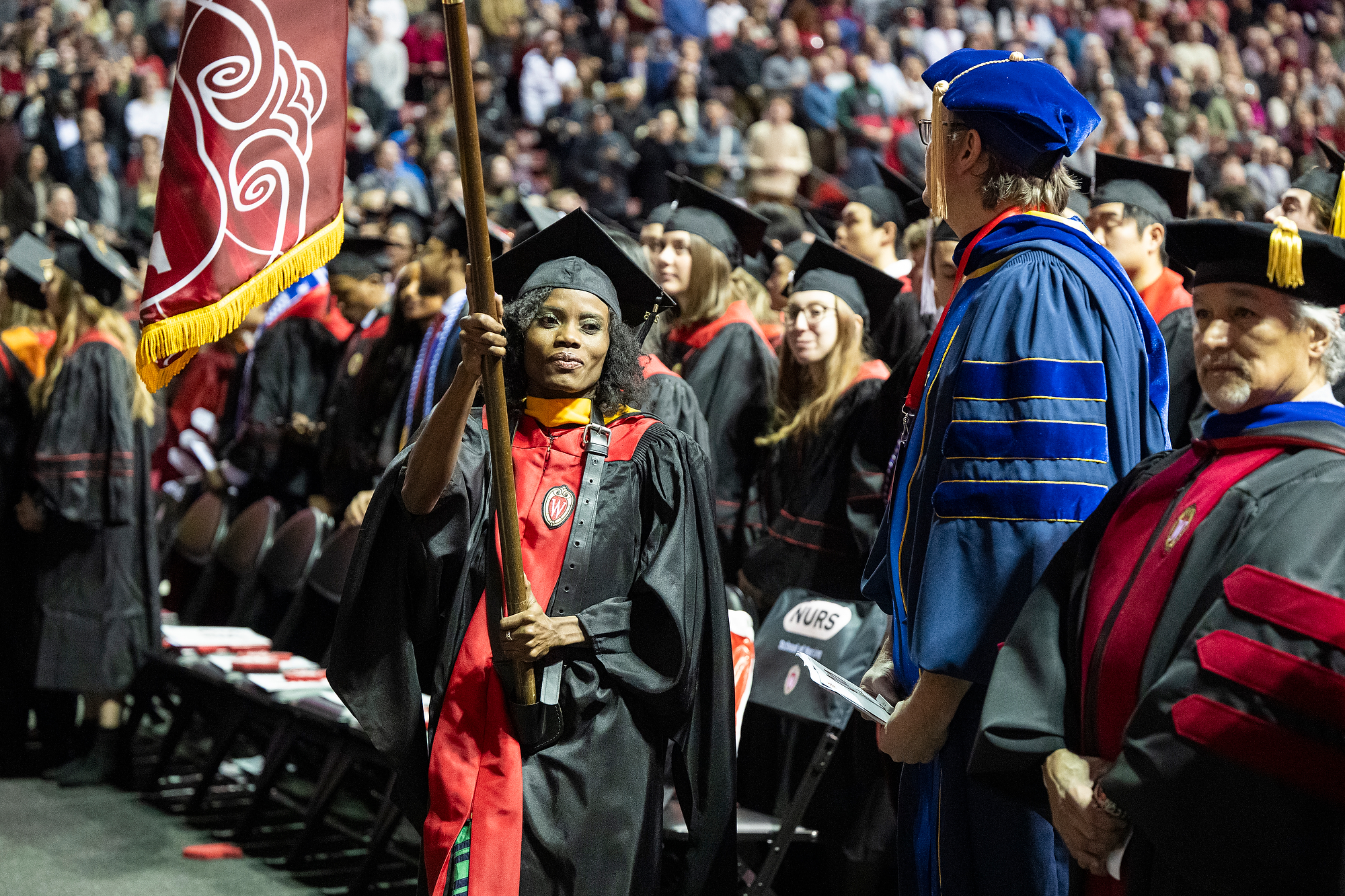 A graduate carries a flag while walking among other graduates and faculty in regalia during a commencement ceremony.