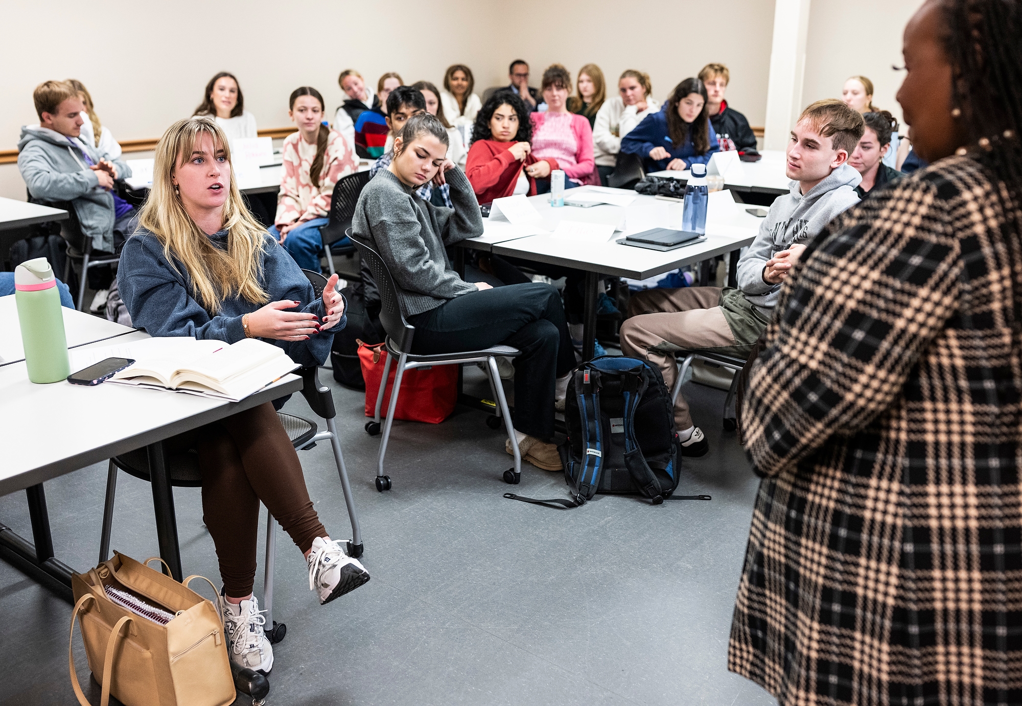 Stubbs stands in the front of a classroom of students seated in at the tables in front of her. She listens to Ellinger who is asking her a question.