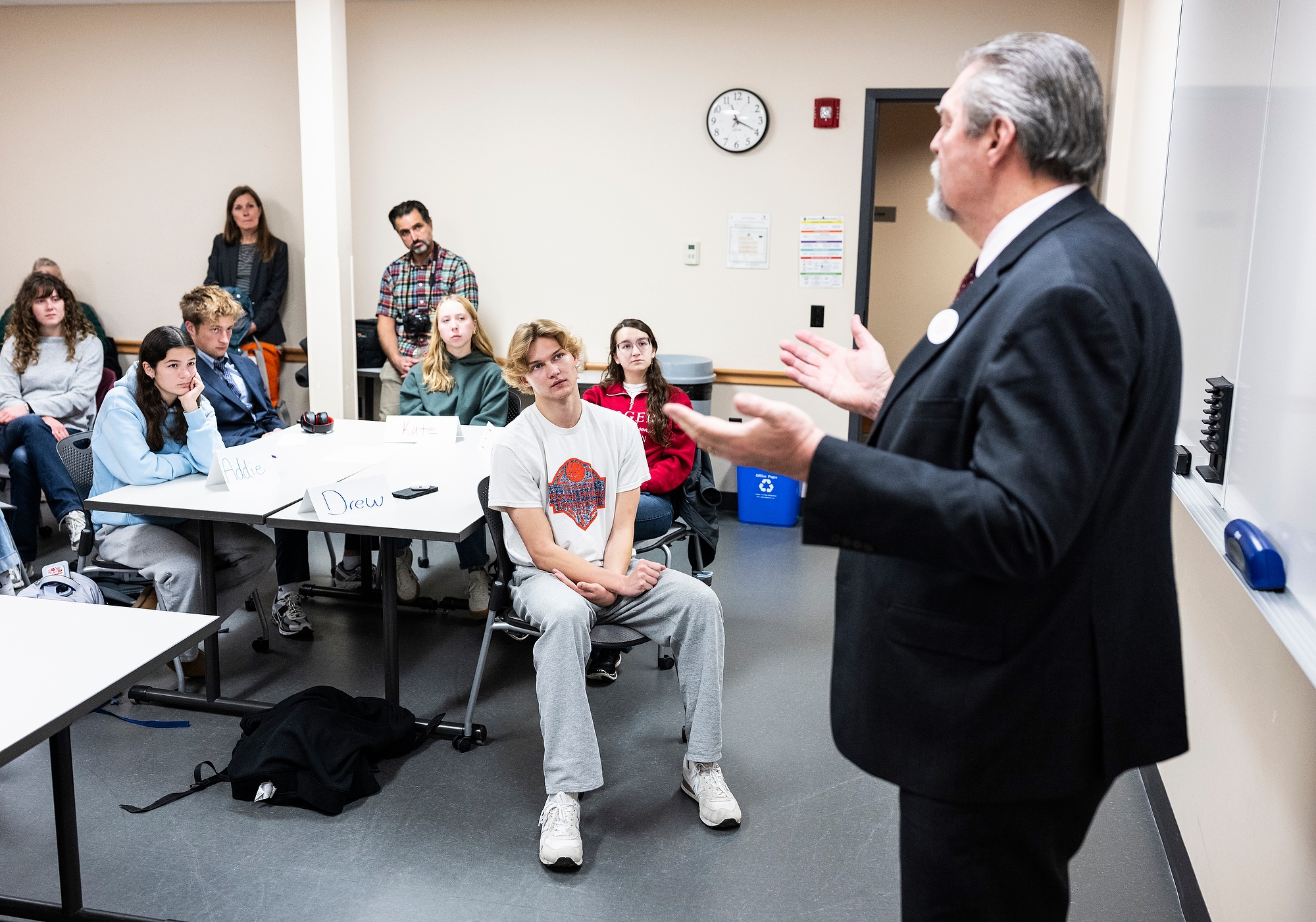 Wittke stands in the front of a classroom addressing students seated in at the tables in front of him.