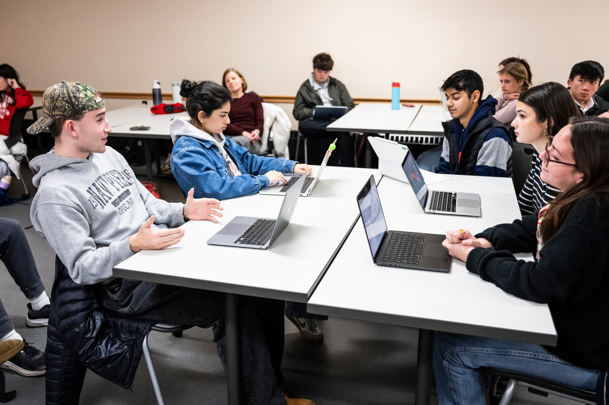 A group of five students sit around a table in a classroom. They each have their laptop open to their notes as the students engage in civil dialogue with one another.