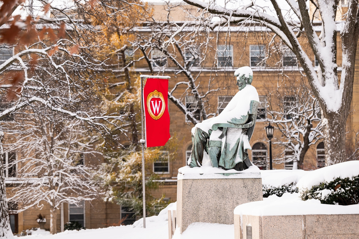 A statue of Abraham Lincoln is covered with snow. Stone buildings and a red banner with the UW "W" logo are seen in the background.