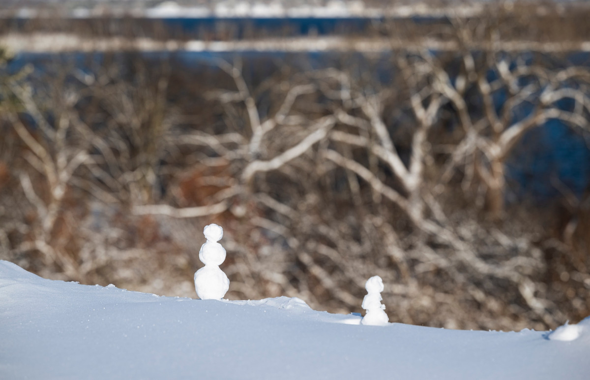 Two tiny snowmen are built on the top of a hill overlooking a lake, so they appear to be looking at the lake.